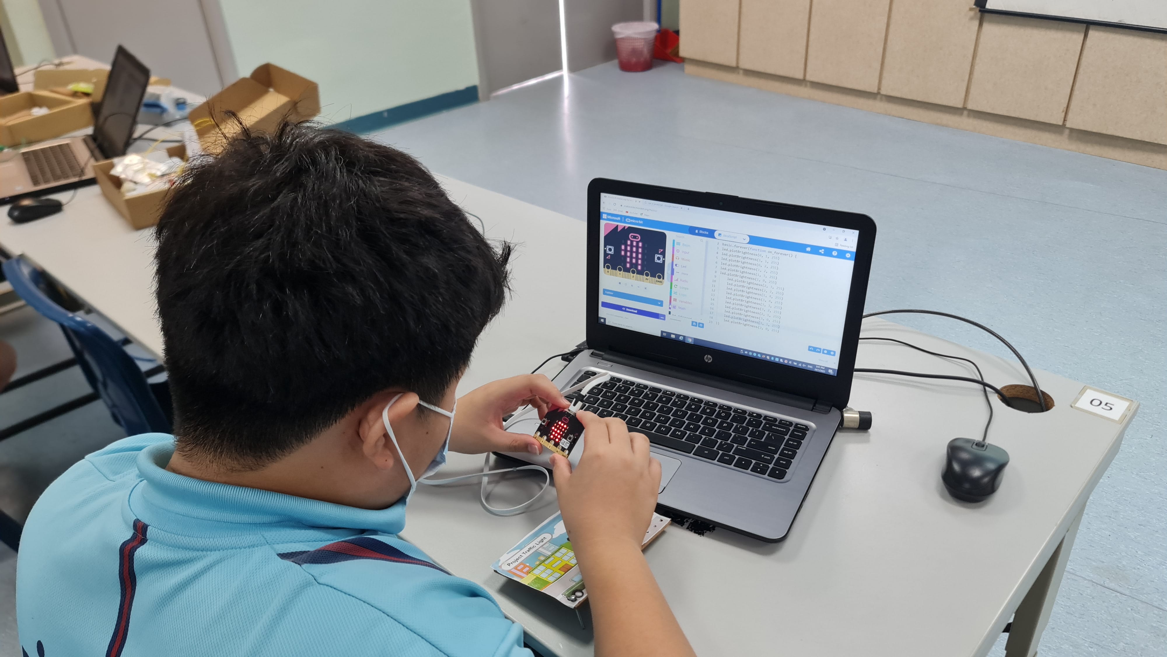 A masked student holds a micro:bit device connected to a laptop showing a MakeCode programming interface.